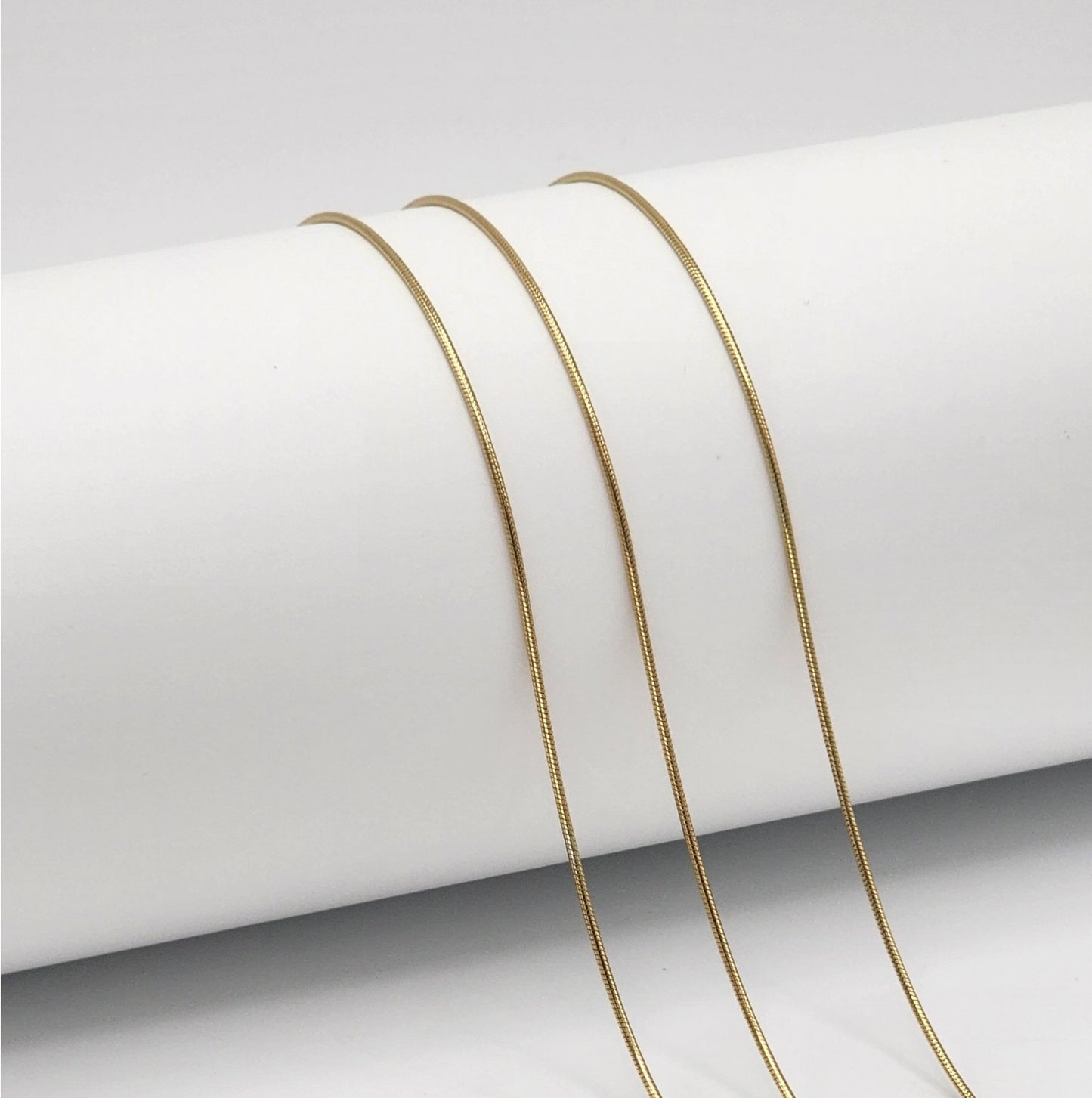 three gold snake chains draped over a white cylinder with a white background.