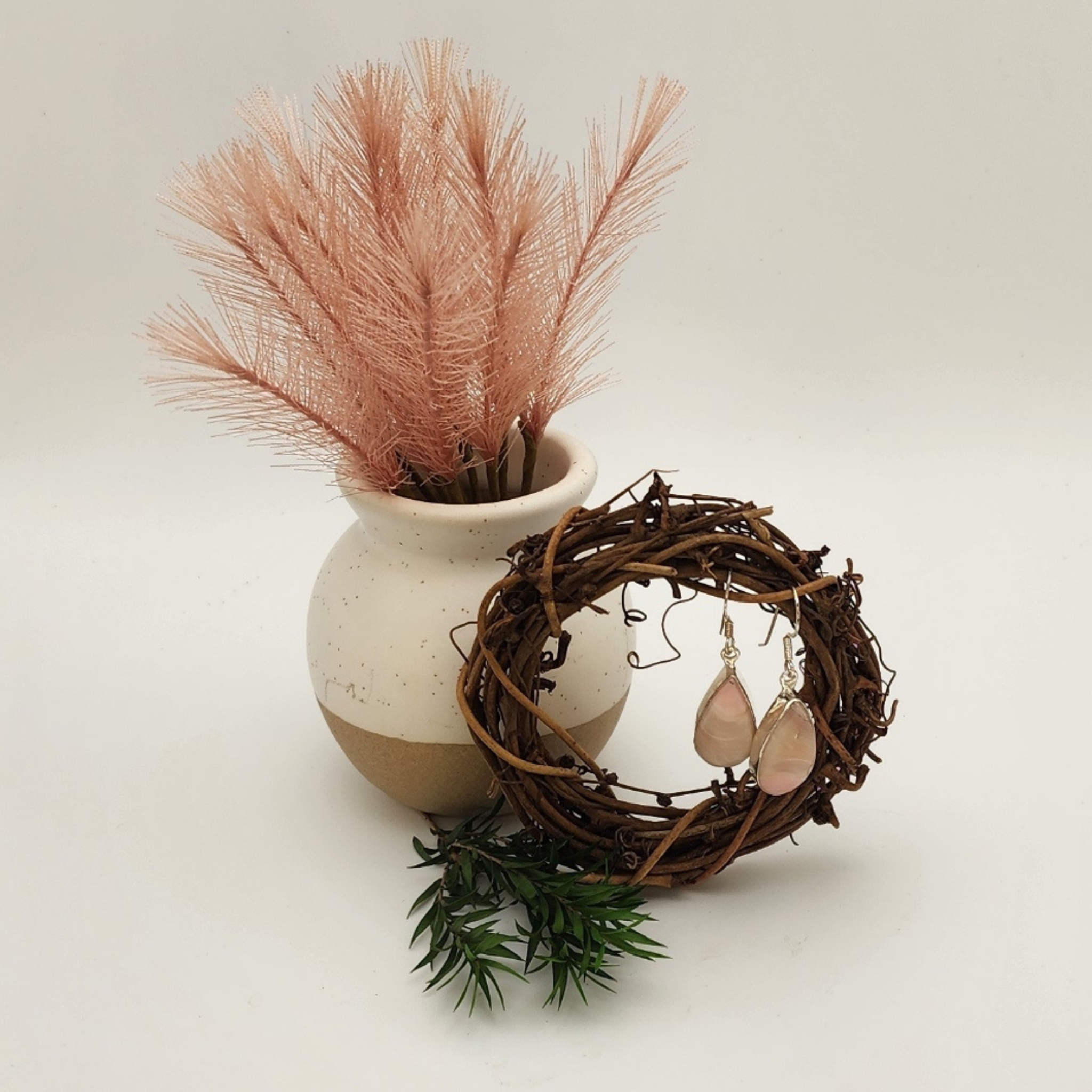 since pair of ice cream opal earrings displayed in a mini wreath made of died brown vines. there is a small pot with a plume of pink feathery stems behind and to the left of the wreath with some greenery below the vase.