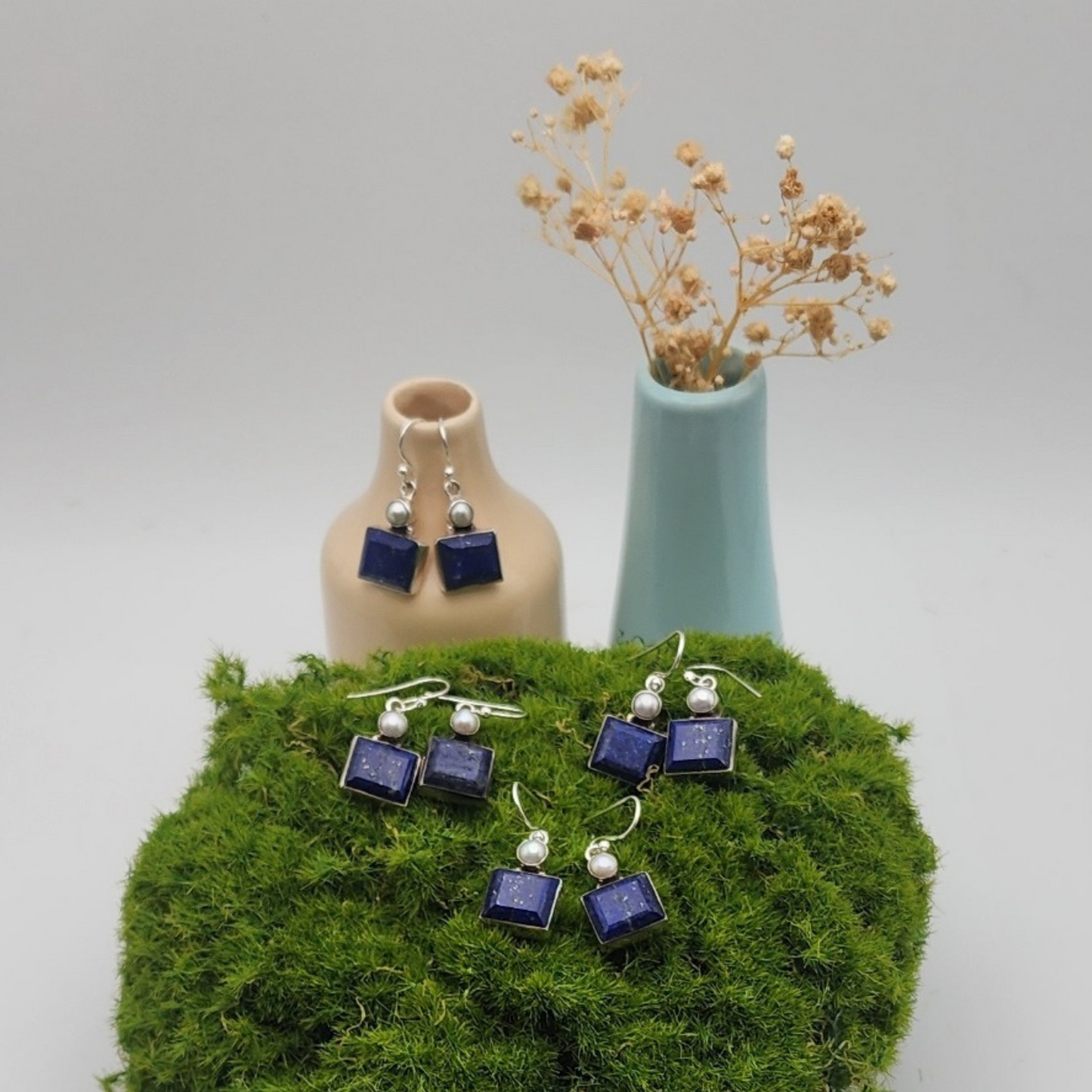 three pairs of lapis and pearl earrings displayed on a piece of moss. behind the moss there is a cream colored vase displaying a pair of lapis and pearl earrings. to the right of the cream vase is a turquoise vase with dried white flowers.