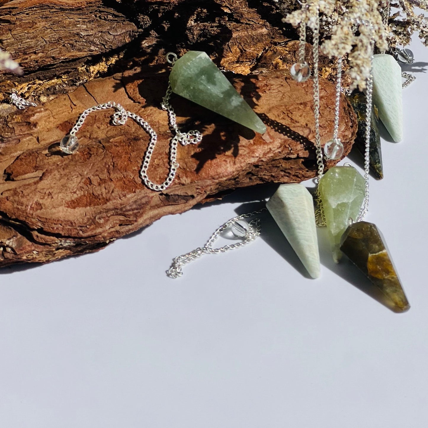 six pendulums with silver chains in varying shades of green stones are draped over a piece of petrified wood