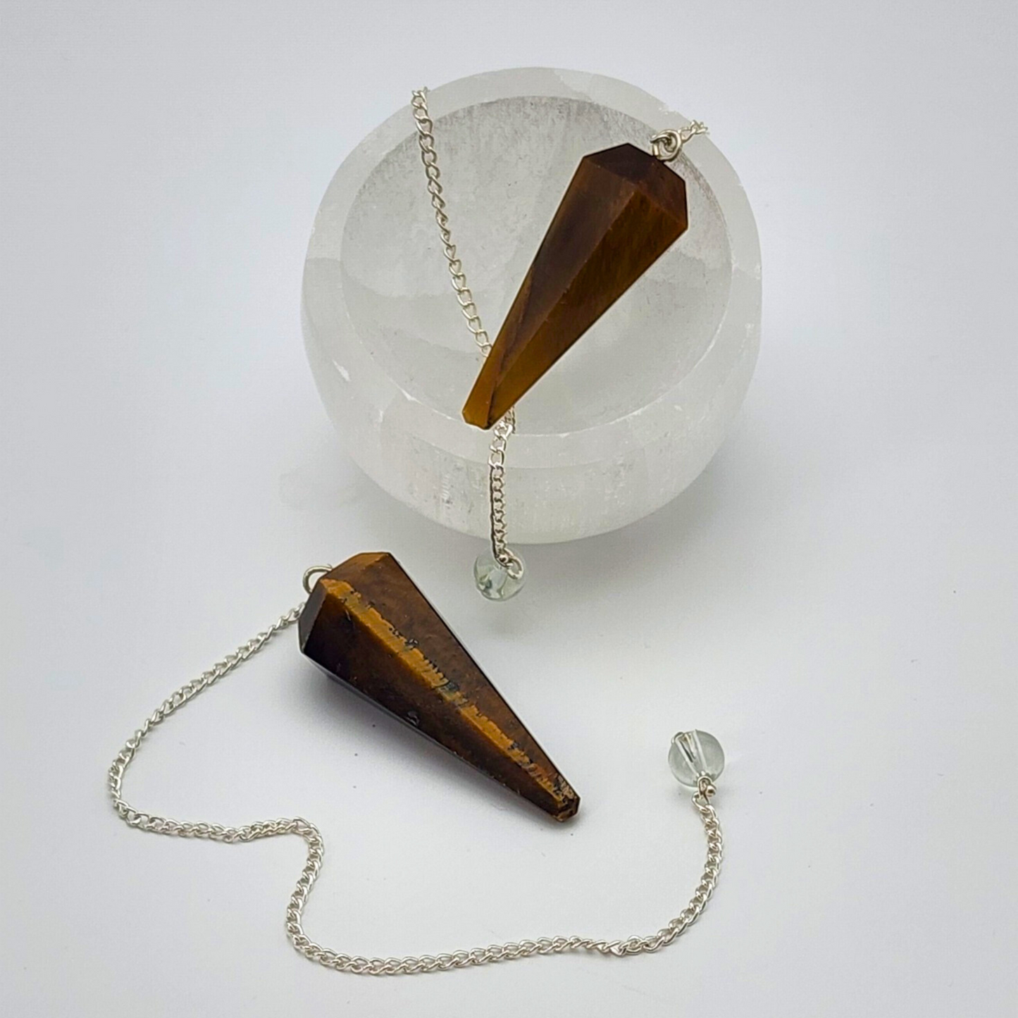 two tigers eye pendulums. one displayed inside of a small selenite bowl with the chain draped over the edge. the other pendulum is in front of the selenite bowl with the chain swirled in front.