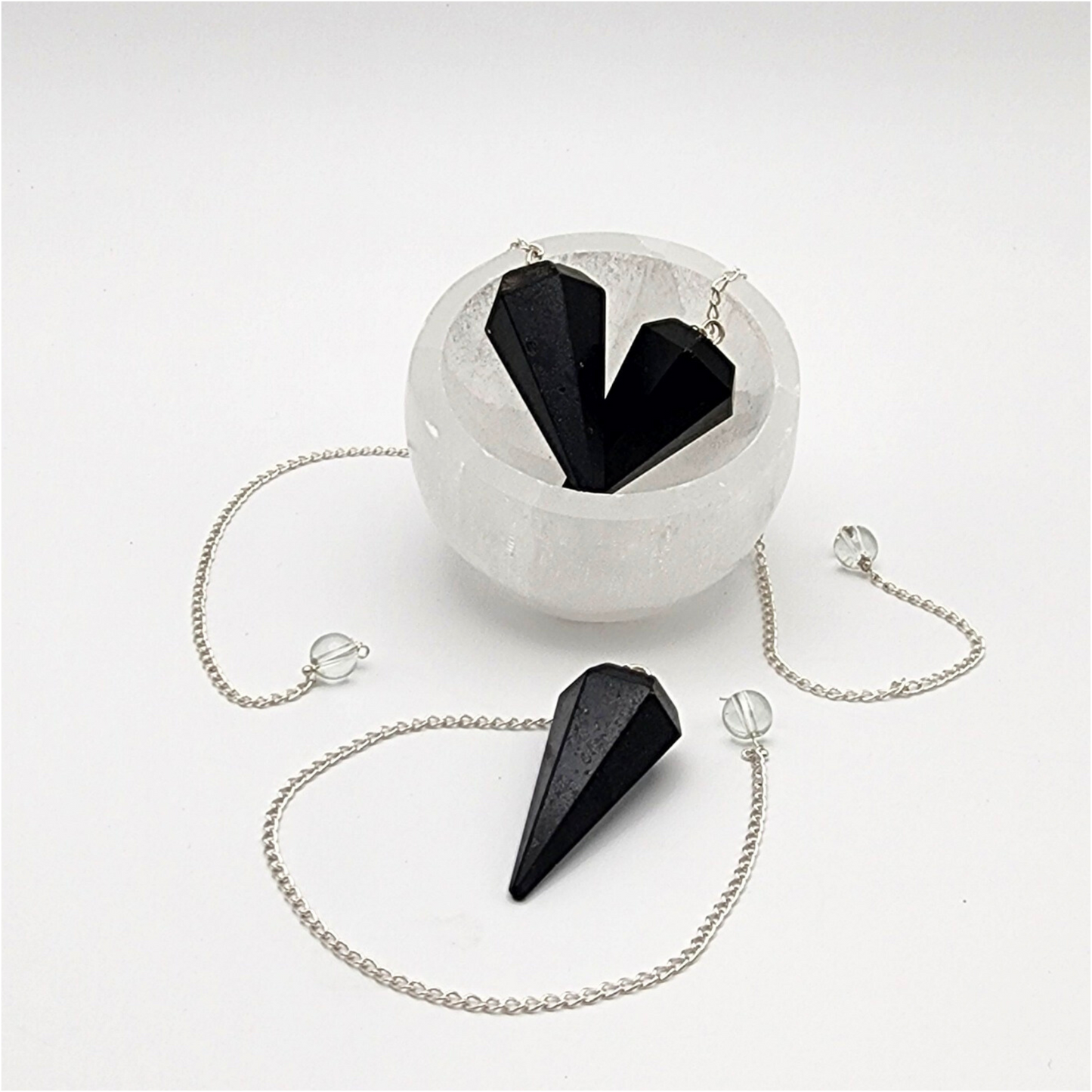 three black tourmaline pendants. two inside a small selenite bowl with the chains draped over the back of the bowl. one outside the bowl with the chain curved around it