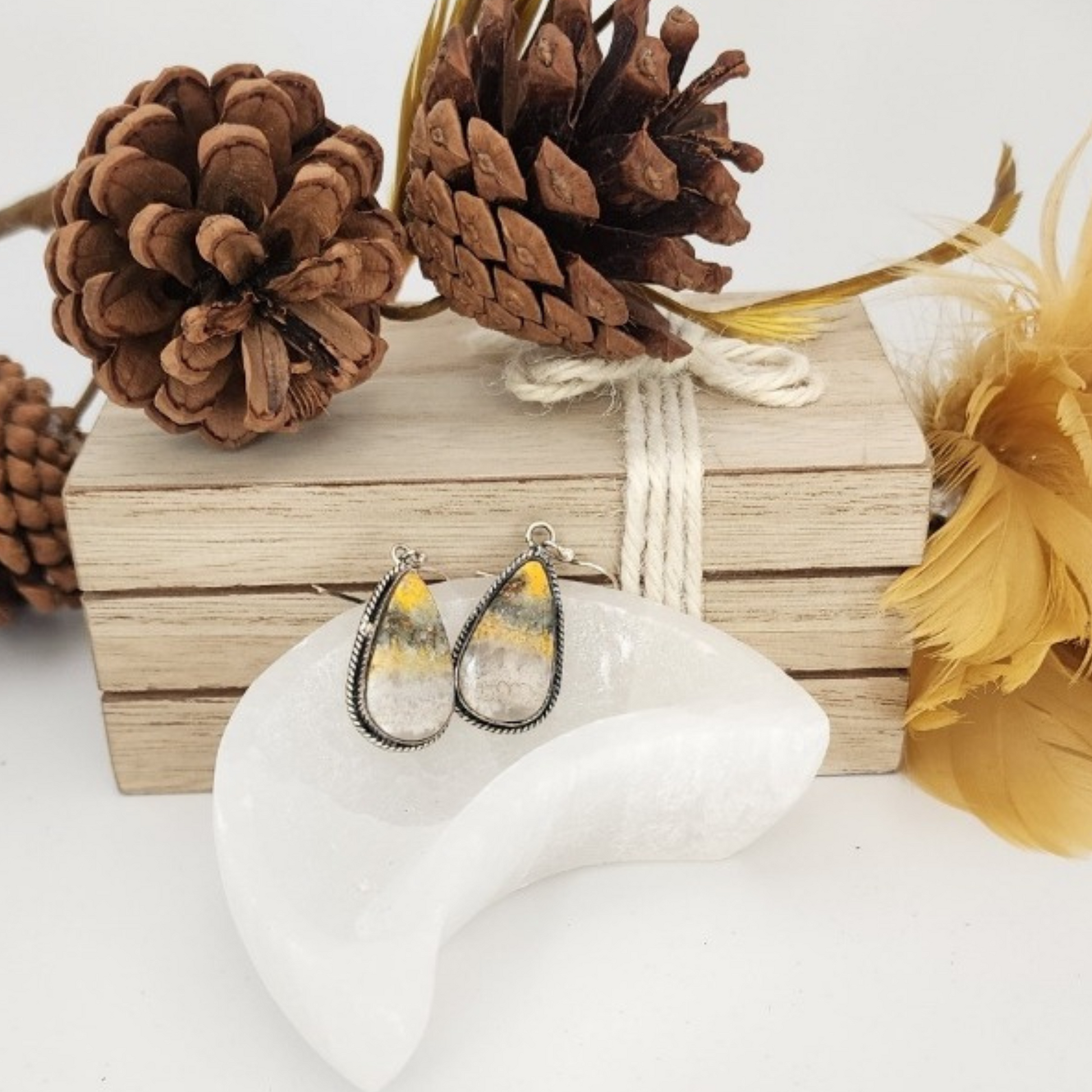 Teardrop-shaped bumblebee jasper earrings in a selenite moon bowl with pinecones and wood in the background