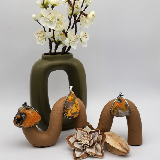three different bumblebee jasper pendants displayed on a terracotta colored squiggle with an olive green vase behind filled with white flowers. two wooden props rested in front of display.