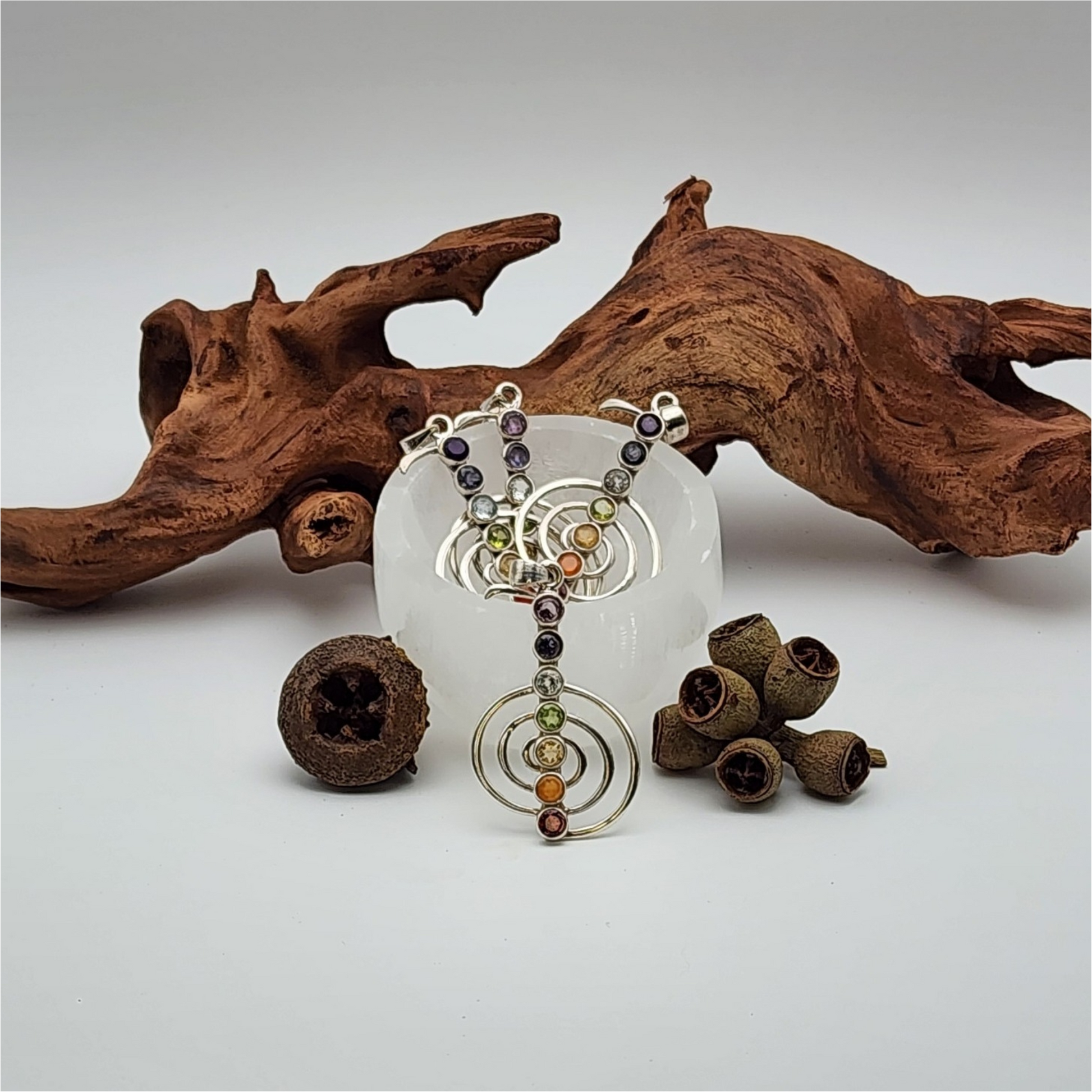 Four chakra pendants in a small selenite bowl with a piece of wood and dried acorns surrounding the bowl.
