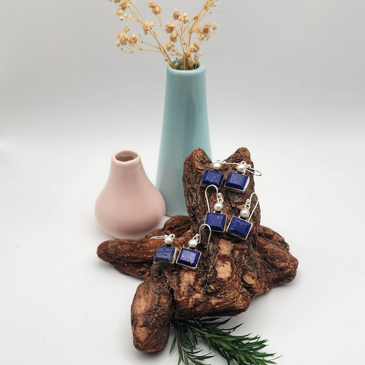 three pairs of lapis and pearl earring displayed on an aged piece of wood. turquoise vase with dried flowers and an empty pink vase are behind the wood. There is a dried piece of greenery below the wood