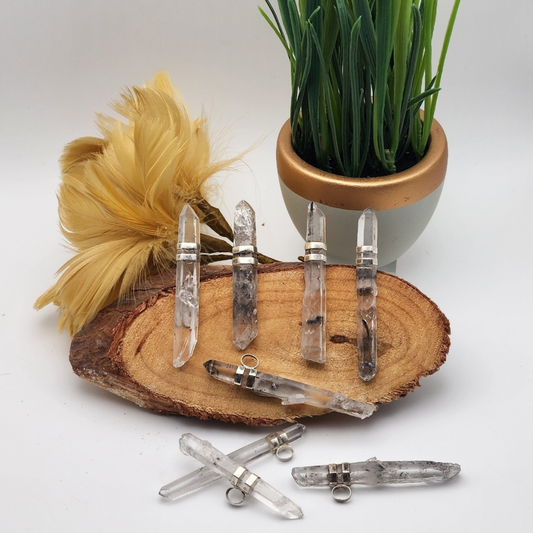 five lemurian quartz pendants displayed on a cross section of wood with three more laid in front of the wood. behind the wood there is a plume of yellow feather to the left and a small pot with grass on the right.