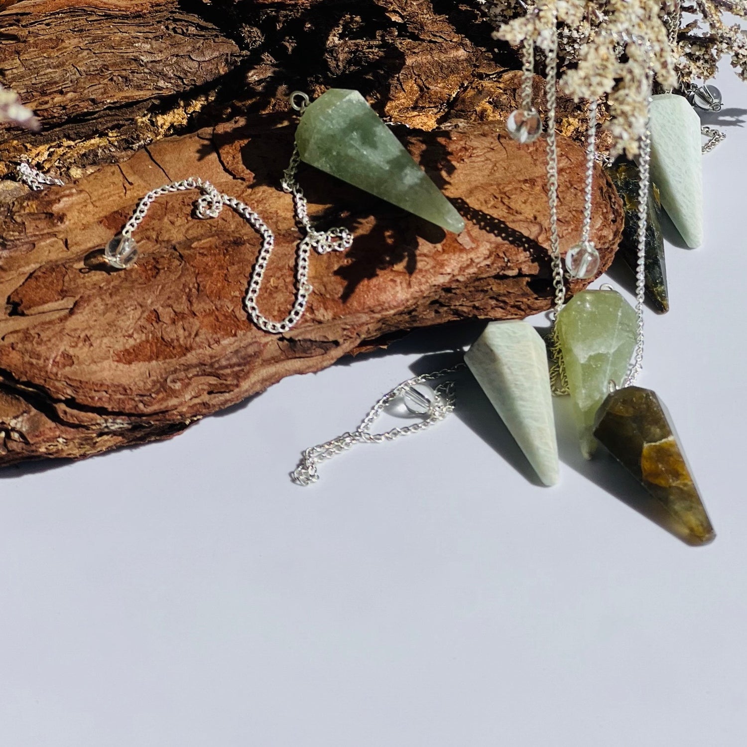 six pendulums with silver chains in varying shades of green stones are draped over a piece of petrified wood
