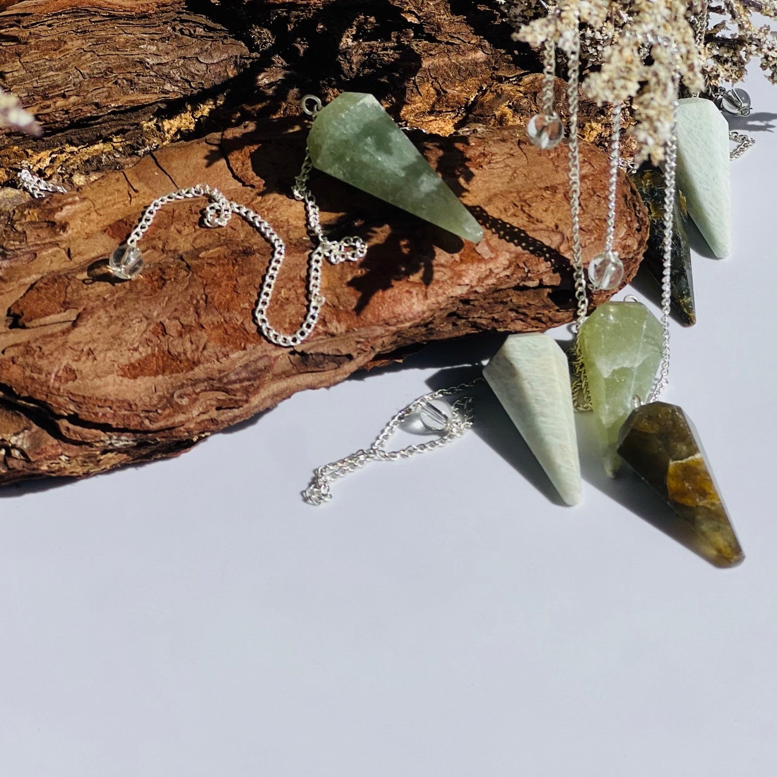 six pendulums with silver chains in varying shades of green stones are draped over a piece of petrified wood