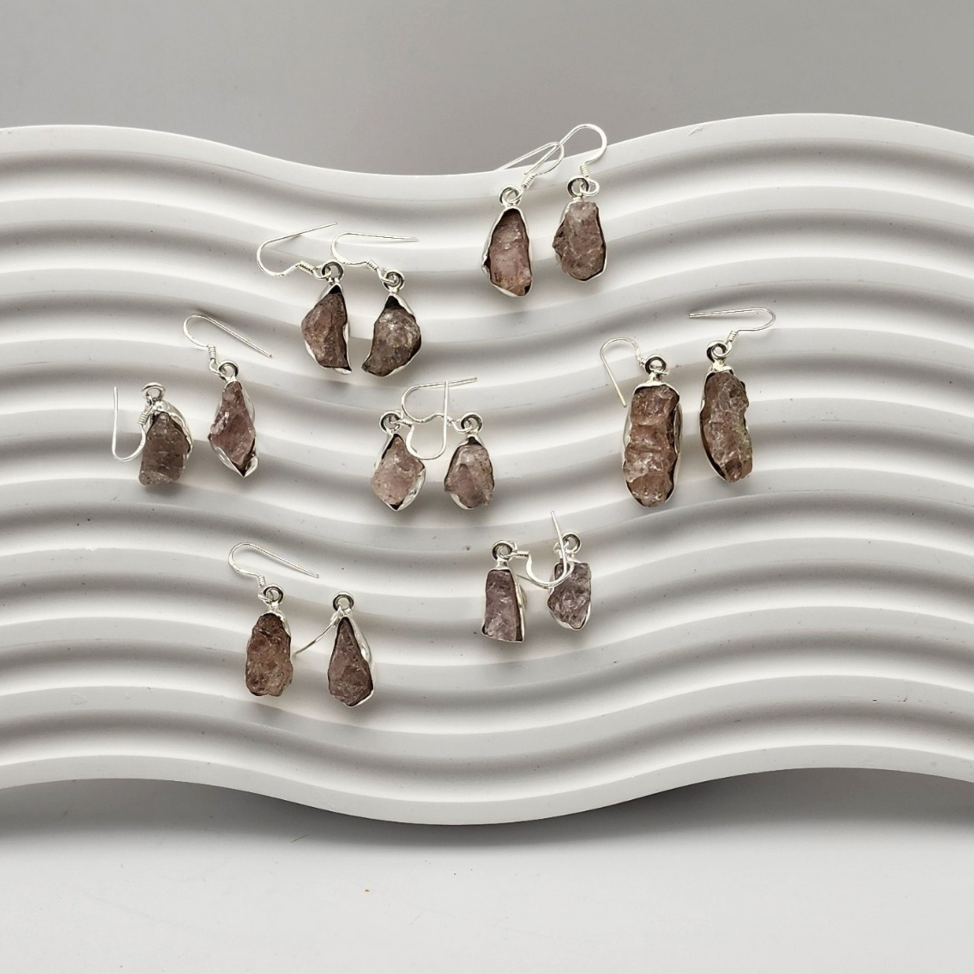 Seven pairs of rough rose quartz earrings laid on a white squiggly background.