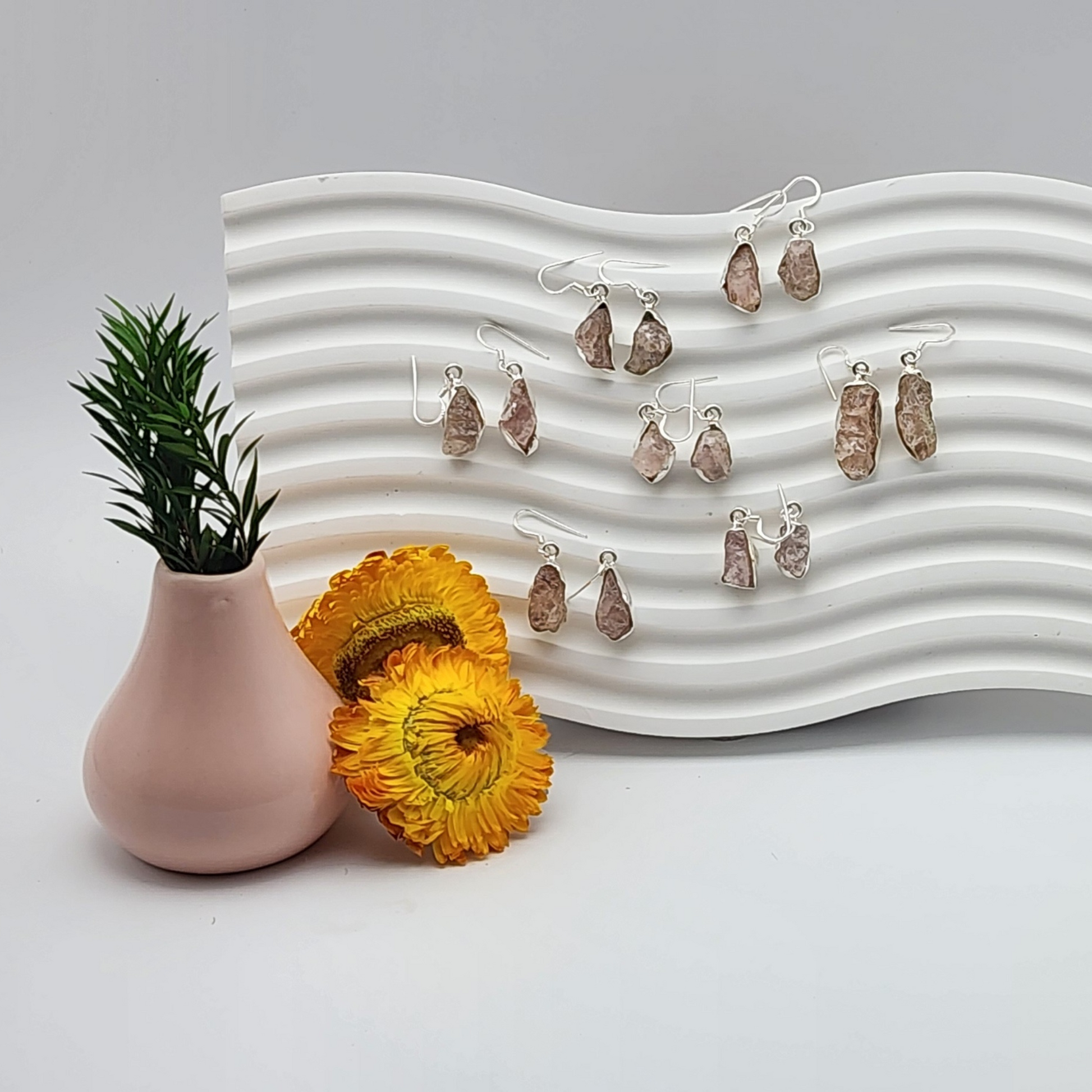 seven pairs of rough rose quartz earrings on a white squiggly background staged and arranged in a moody Bloom and Glow aesthetic scene.