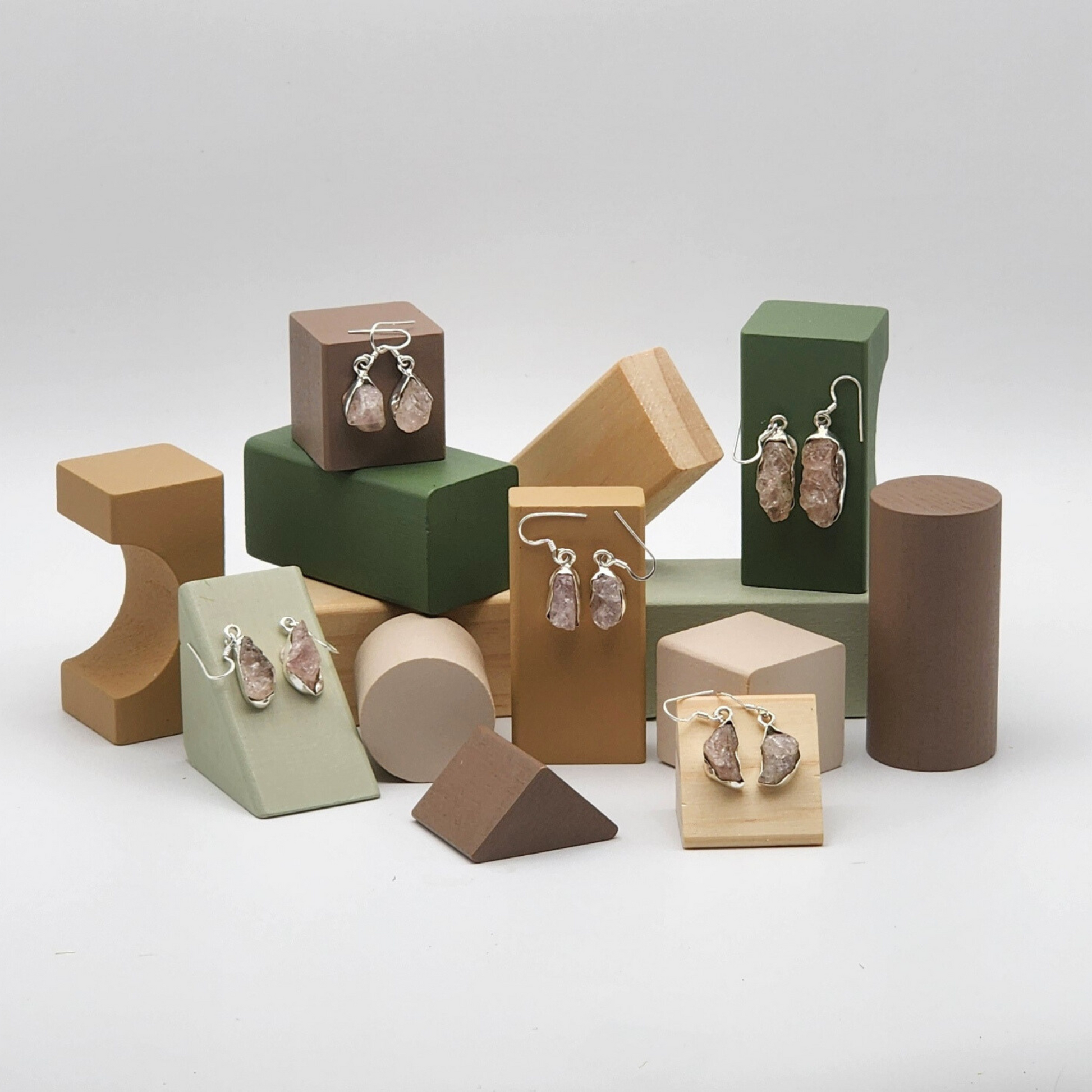 five pairs of rough rose quartz earrings displayed on assorted colored blocks. blocks are various neutral tones and various different shapes.