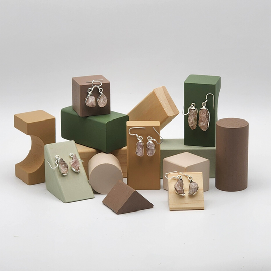 five pairs of rough rose quartz earrings displayed on assorted colored blocks. blocks are various neutral tones and various different shapes.