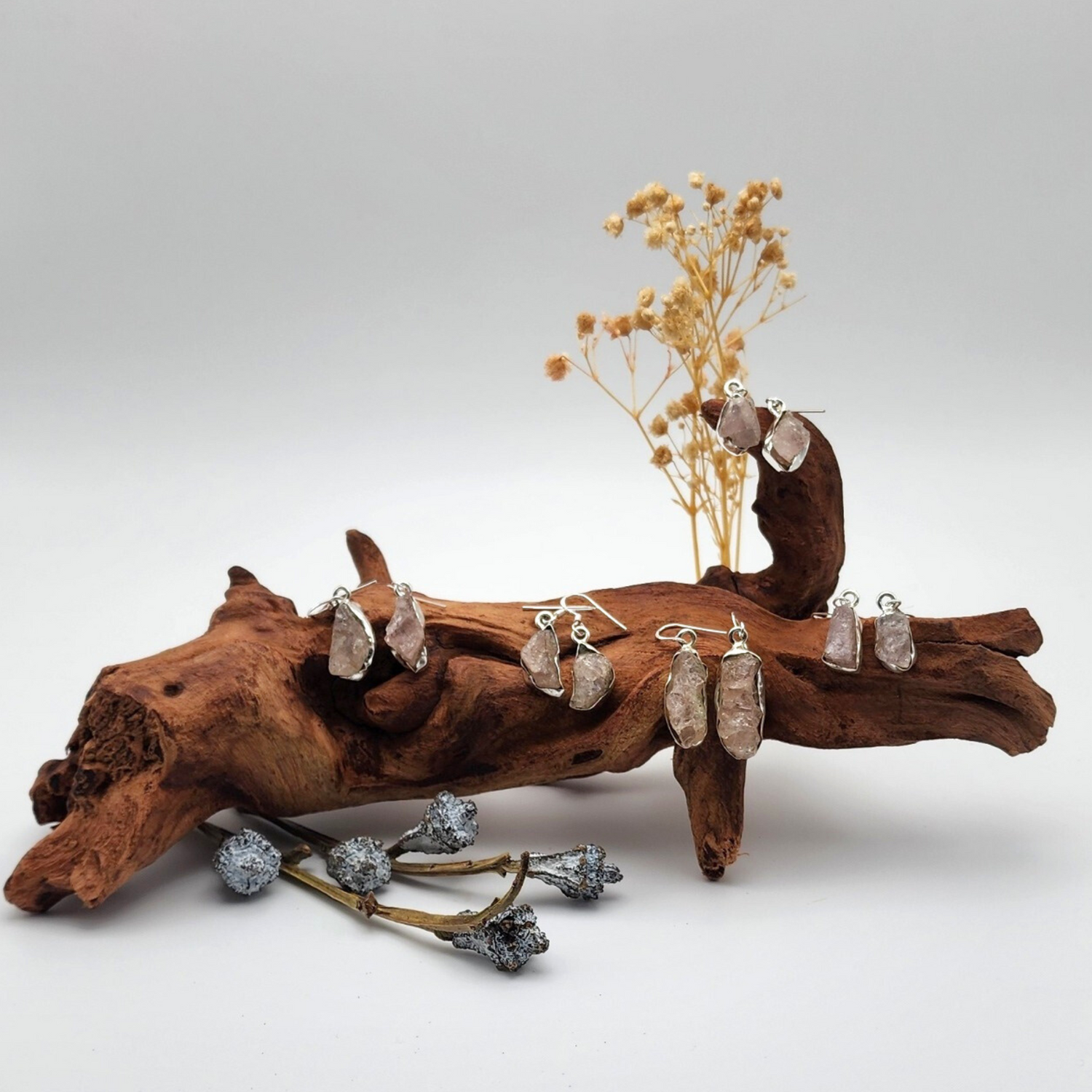 five pairs of rough rose quartz earrings displayed on a piece of dried wood with dried florals behind and in front of the wood.