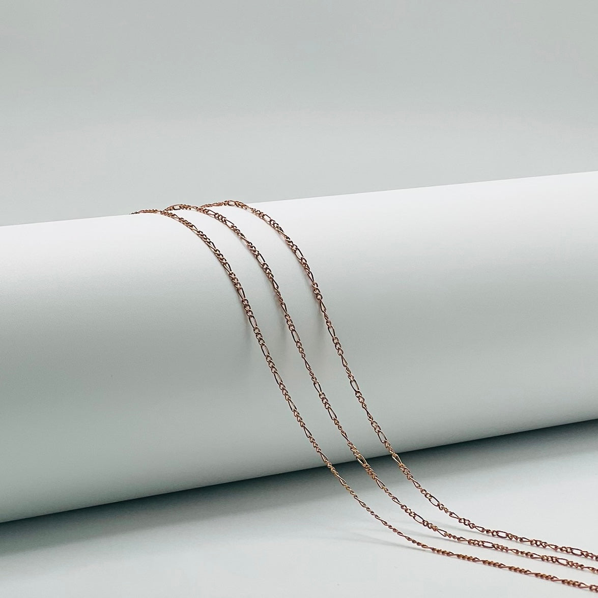 three rose gold figaro chains draped over a white cylinder with a white background.