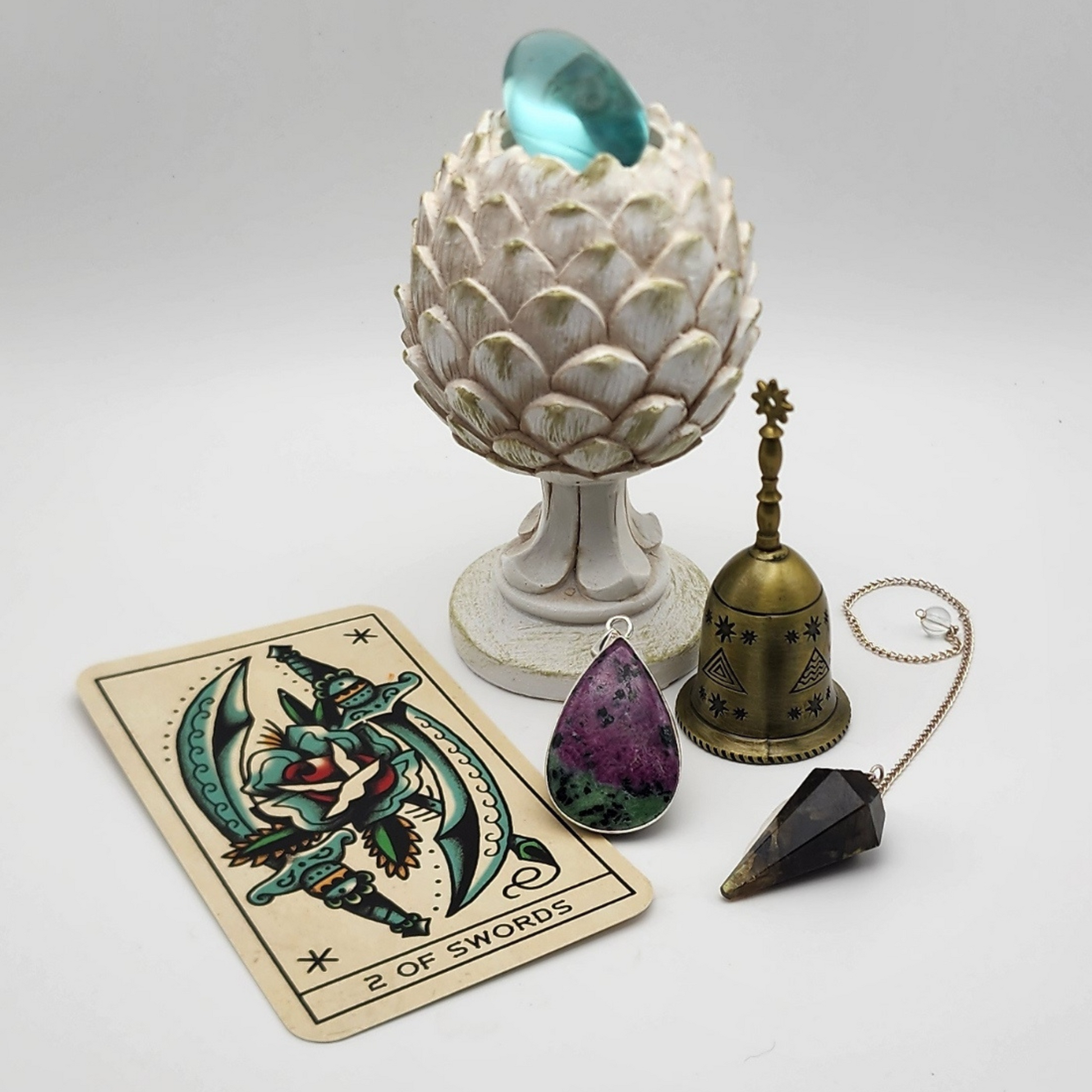 single ruby zoisite pendant set in a display scene with a two of swords tarot card to the left, a brass bell and labradorite pendulum to the right staged and arranged in a moody Bloom and Glow aesthetic scene.