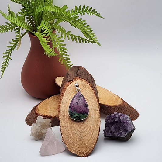 single zoisite pendant displayed on a sliced cross section of wood. there are two other slices of wood behind the display and a terracotta colored vase with greenery. there are two raw crystals on the right and a single raw crystal to the left of the displayed pendant.
