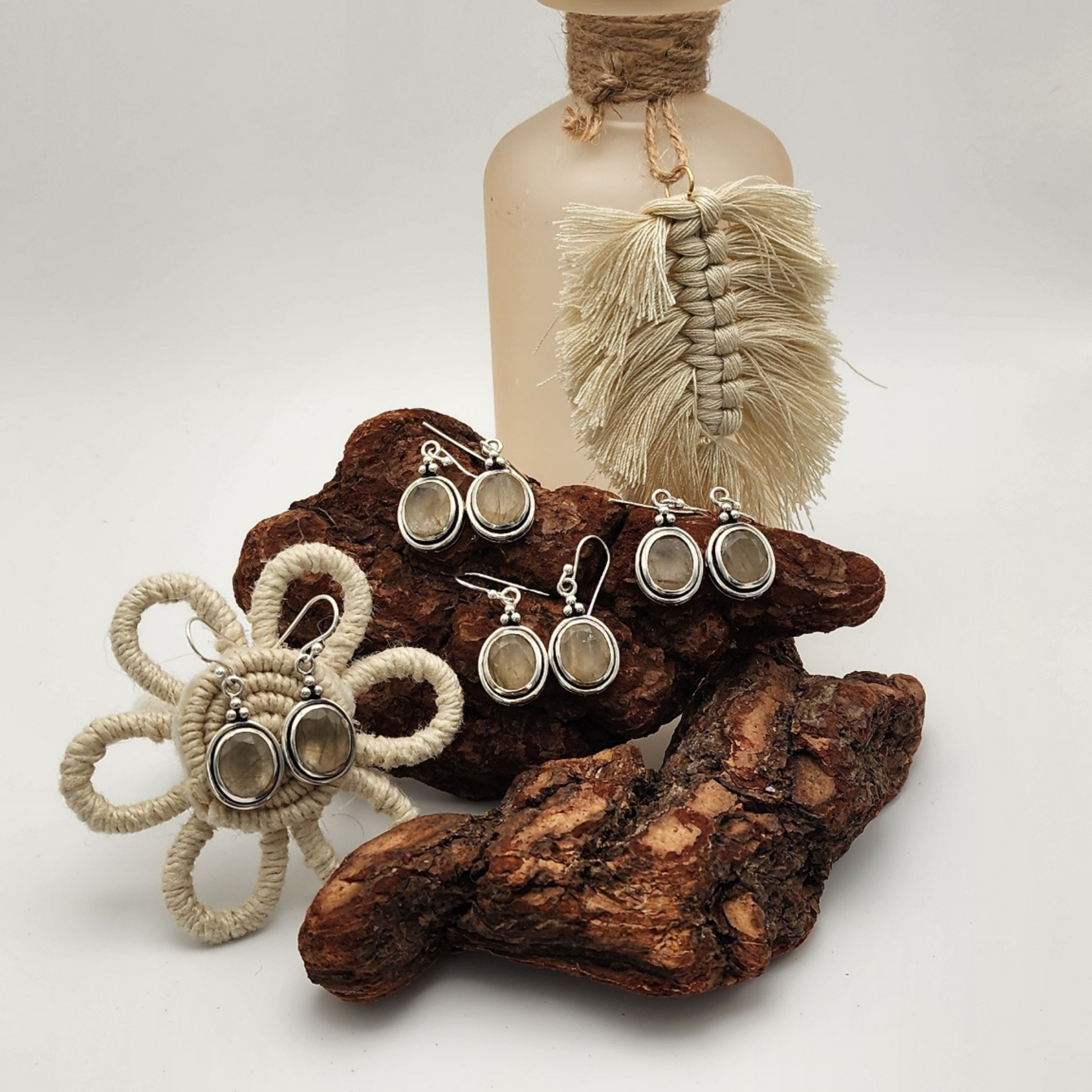 four pairs of faceted rutilated quartz earrings three are displayed on an aged piece of wood with another pair displayed on a flower shape made of beige rope. There is a frosted pink vase with decorative tassel hanging off the vase.