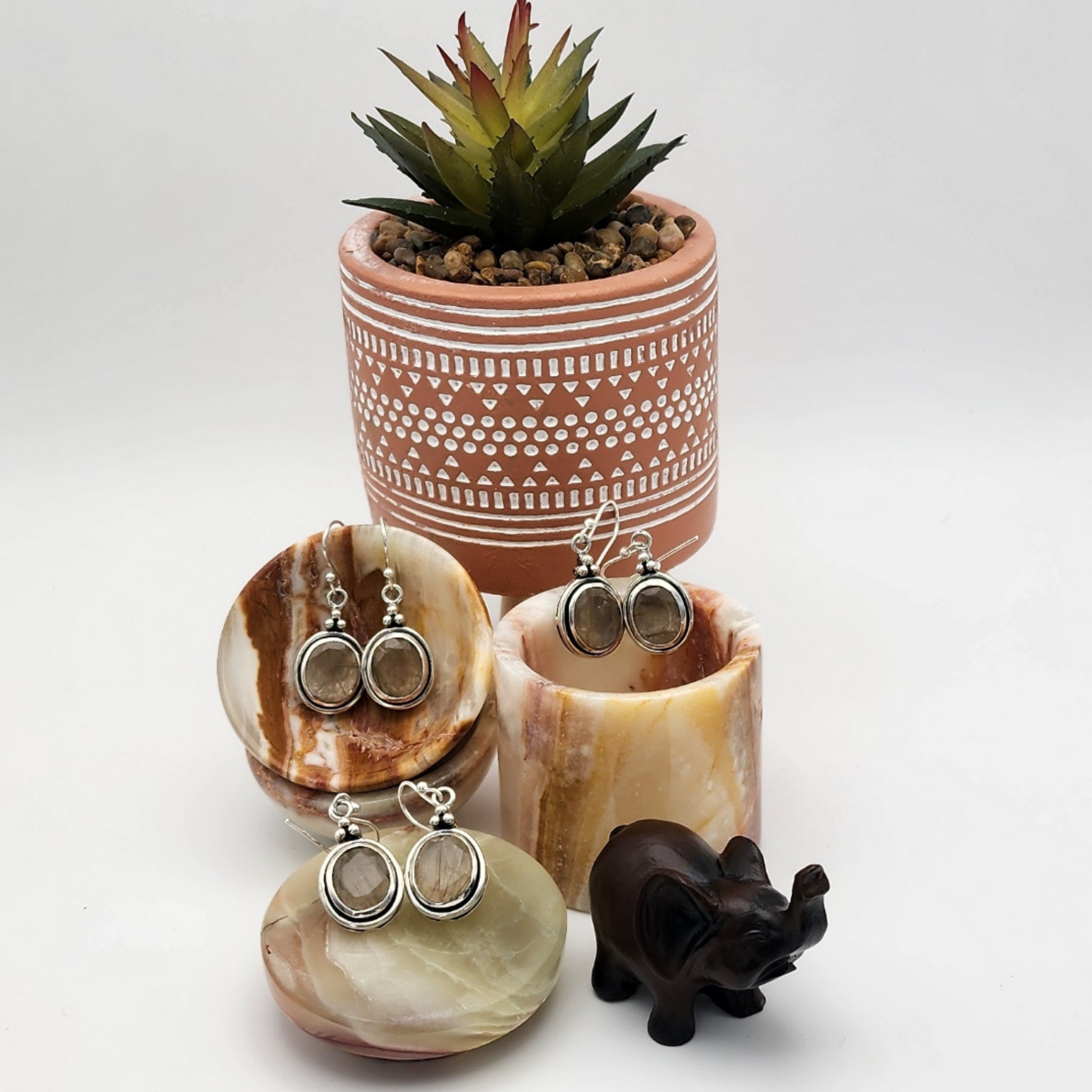 three pairs of faceted rutilated quartz earrings displayed in a scene with two small onyx trinket bowls and a small onyx jewelry box with lid. one pair is inside the trinket bowl, another pair inside the jewelry box, another pair on the box lid. There is a potted succulent with gravel behind the onyx and a small wooden cared elephant figurine in front of the jewelry box.