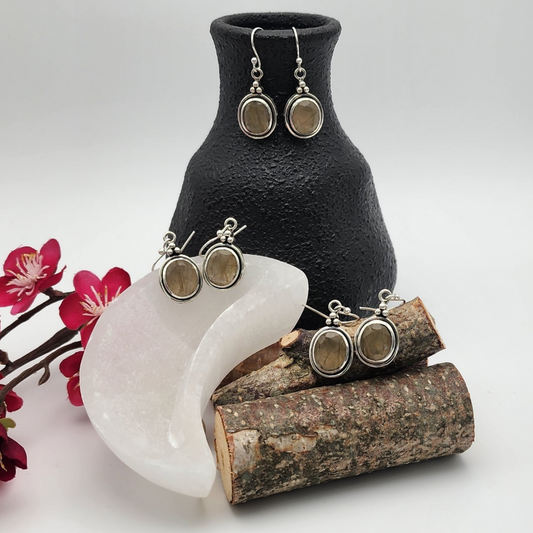 three paired of faceted rutilated quartz earrings displayed within a scene where one pair is in a small black vase, another pair inside a selenite moon trinket dish, and another pair on a mini birch log pile with magenta flowers off to the left behind the selenite dish.