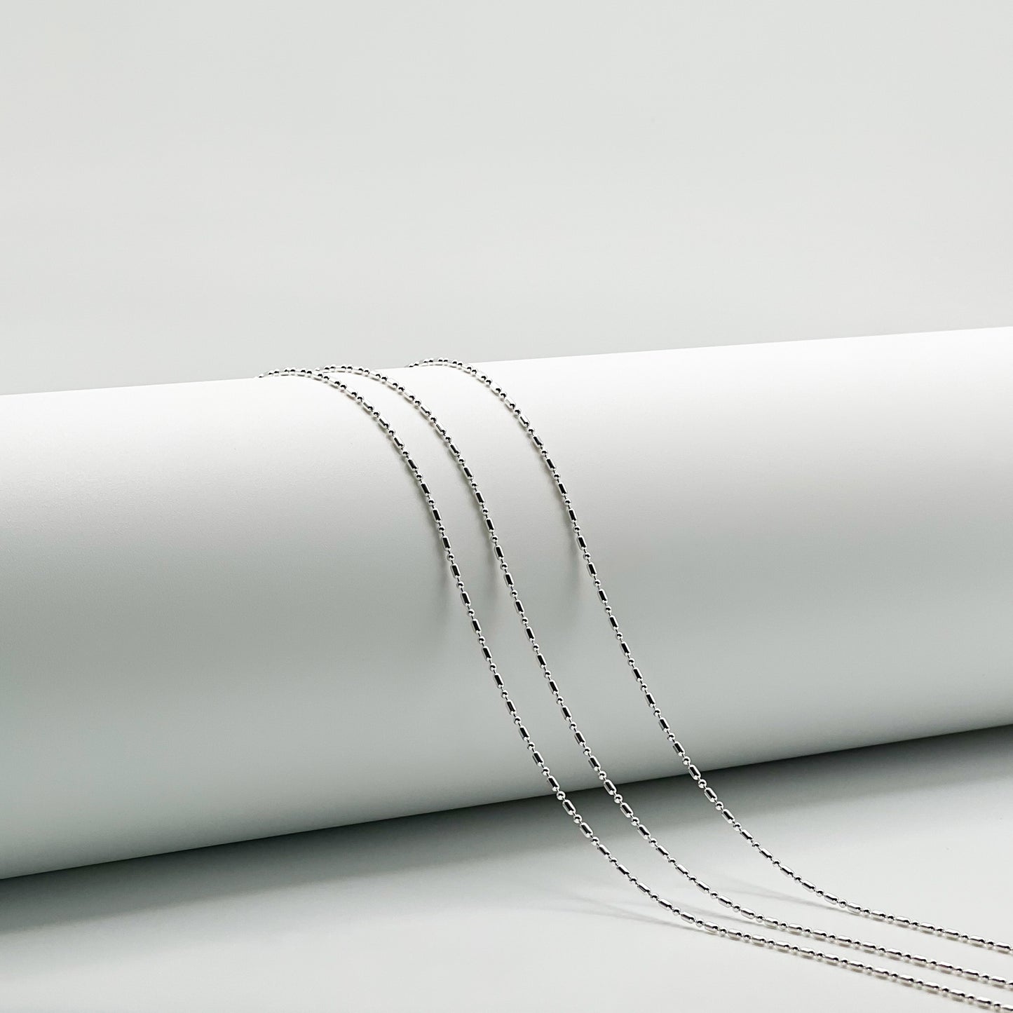 Three silver 1+1 bead necklaces draped over a white cylinder with a white background. 