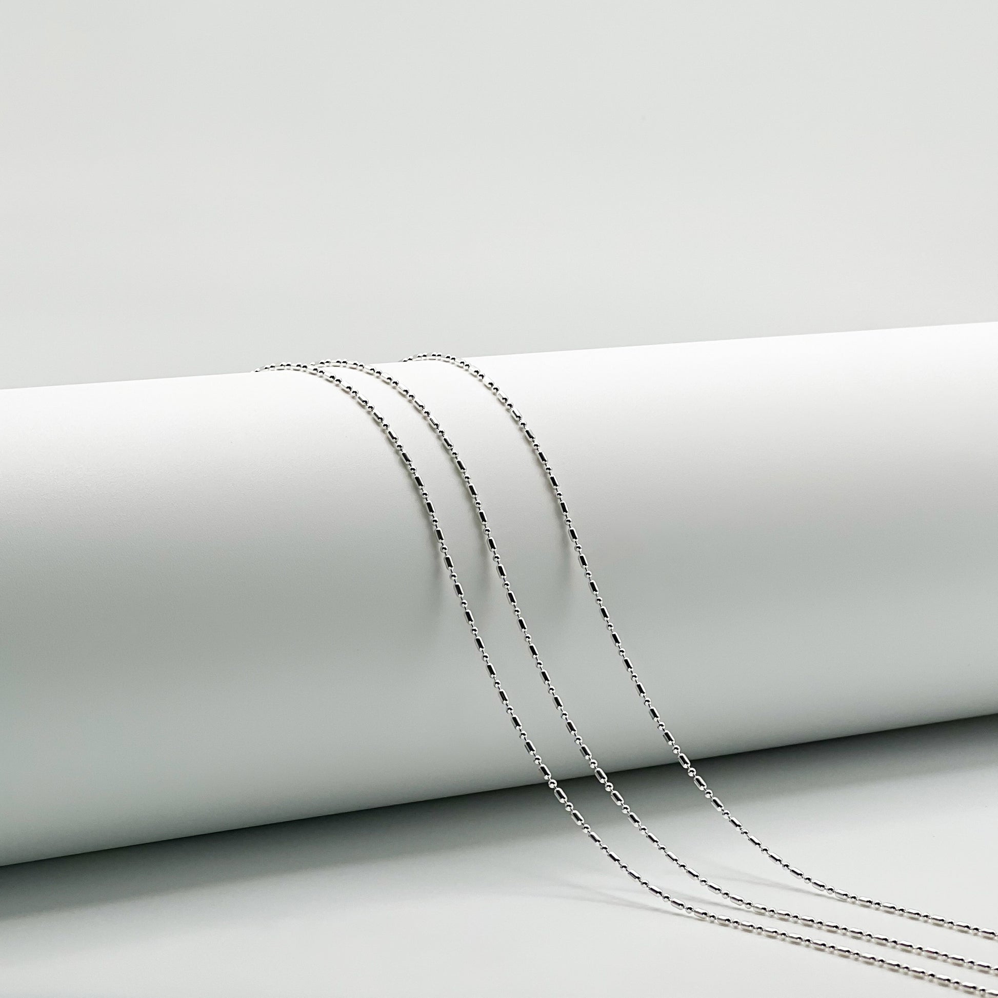 Three silver 1+1 bead necklaces draped over a white cylinder with a white background. 